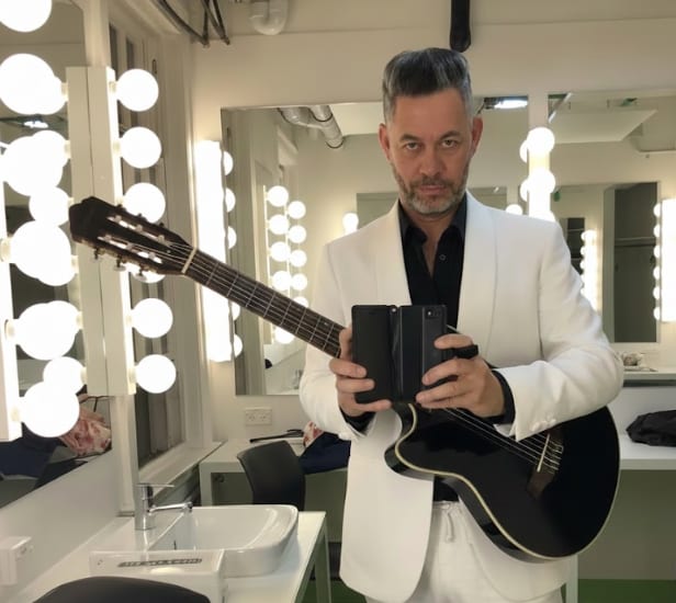 Clinton Mann in a white suit holding a guitar in a dressing room lined with mirror lights
