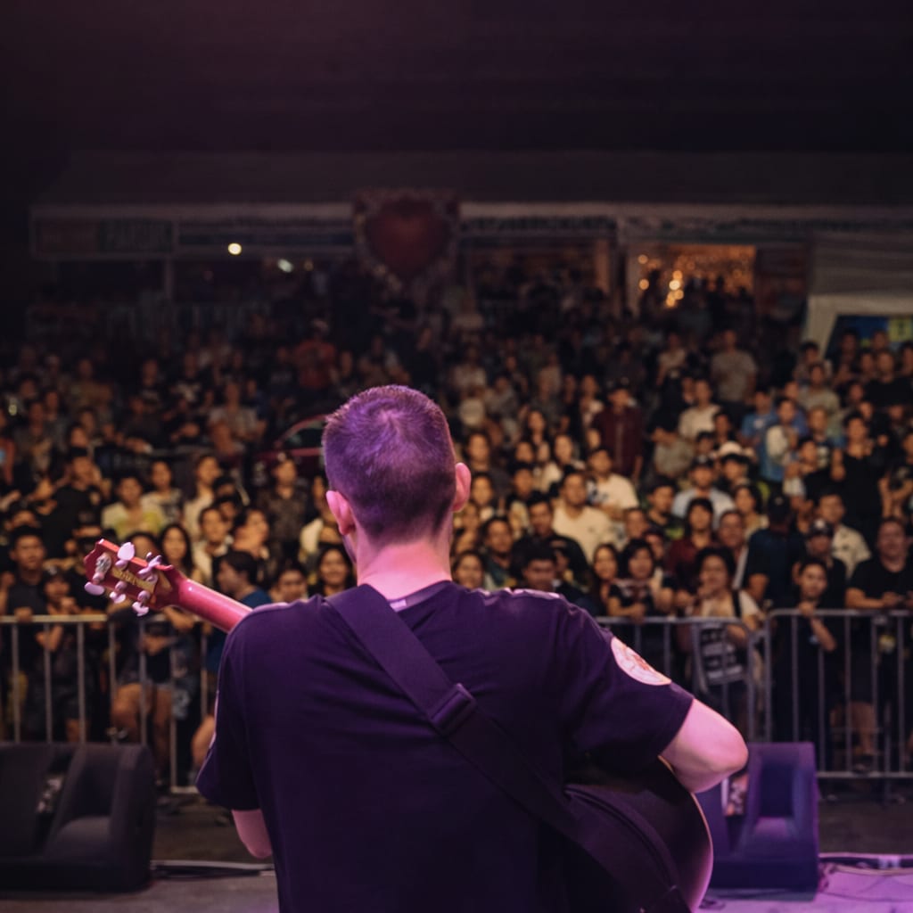 Clinton Mann seen from behind on stage with an acoustic guitar, facing a packed crowd